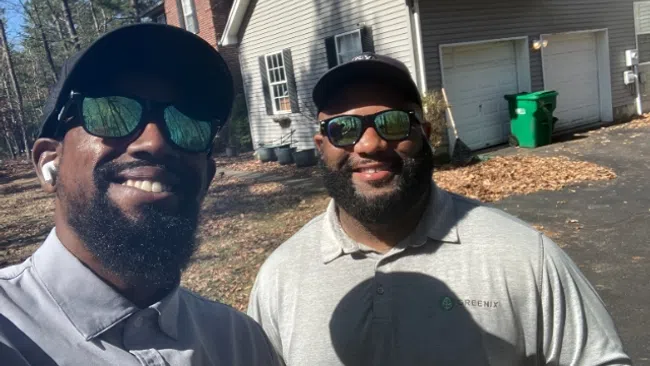 Two men wearing sunglasses and matching Greenix shirts smiling outdoors near a driveway and house.