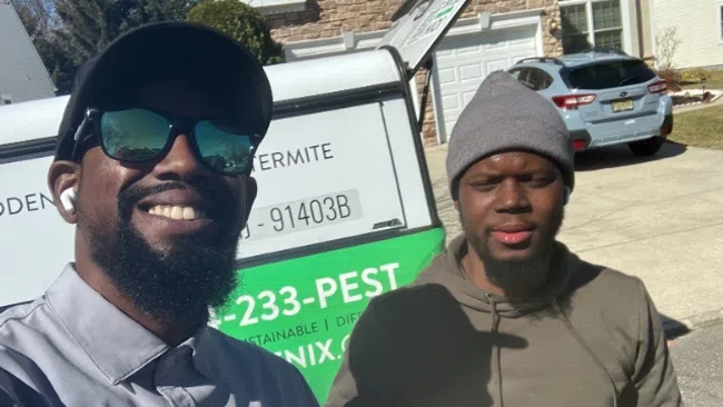 Two men smiling outdoors in front of a pest control service truck on a sunny day in a residential neighborhood