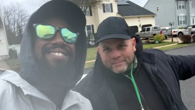 Two men wearing jackets and caps take a selfie outside in a suburban neighborhood on a cloudy day.