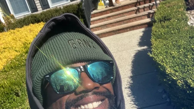 Man in black jacket and green beanie smiling outdoors with reflected trees in sunglasses and house in background.