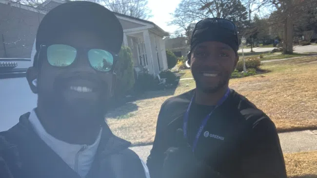 Two men smiling outdoors in a residential neighborhood on a sunny day, one wearing sunglasses and the other giving a thumbs up.
