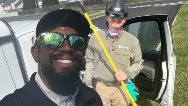 Two men outdoors near a white van smiling, one holding a yellow broom, both wearing sunglasses and hats on a sunny day