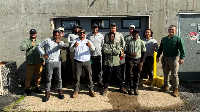 Group of eleven men posing outdoors in front of a concrete building on a sunny day.