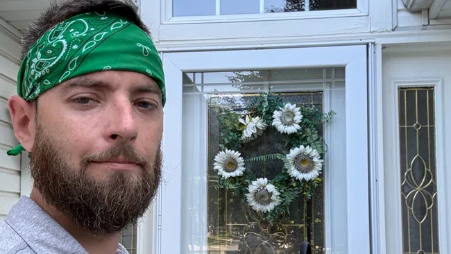 Man with green bandana and beard stands outside white house holding paper in front of decorative glass door with flower wreath.