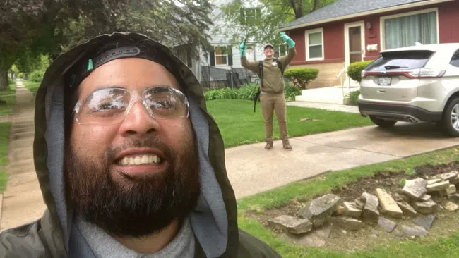 Two men outdoors on a residential sidewalk, one smiling close to camera, the other waving in the background near houses.