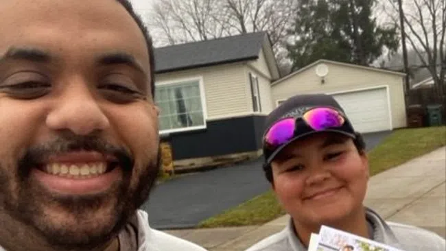 Two smiling people outdoors wearing Greenix jackets, one holding a Greenix-branded information sheet on residential property.