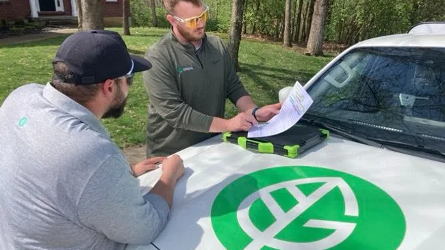 Two men discuss documents on the hood of a white vehicle with a large green leaf logo outdoors.