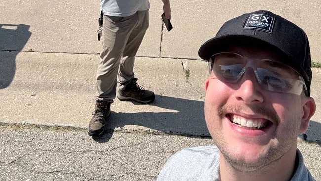 Two men in work attire smiling and posing in front of a suburban house driveway on a sunny day.