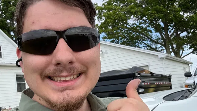 Young man wearing sunglasses and green shirt giving thumbs up outdoors with trees and white buildings behind him