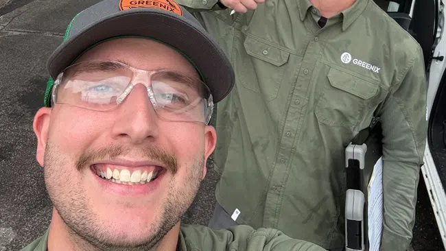 Two smiling landscapers in green uniforms and safety glasses pose for a selfie on a residential street.