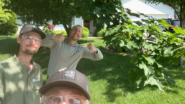 Three landscapers wearing safety glasses and caps posing outdoors in front of a house with green lawn and trees.