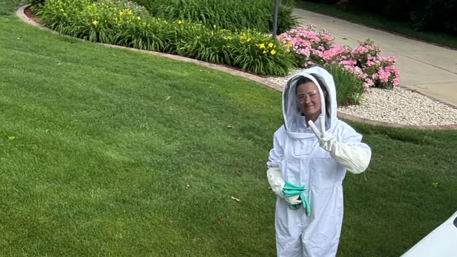 Two people in white beekeeping suits smiling near a white van and green lawn with flowers.