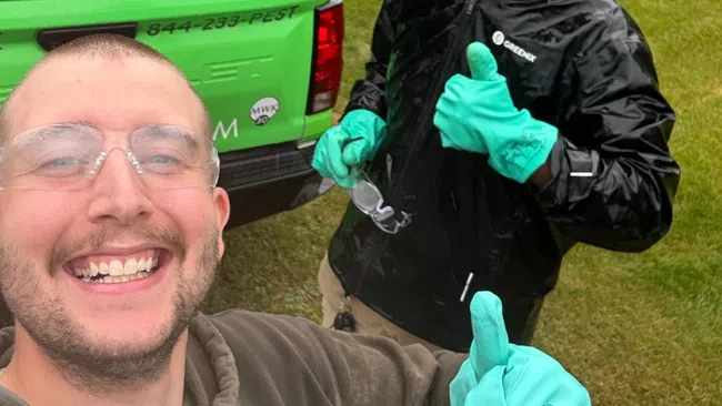 Two pest control workers in protective gloves giving thumbs up next to a green truck on a grassy lawn.