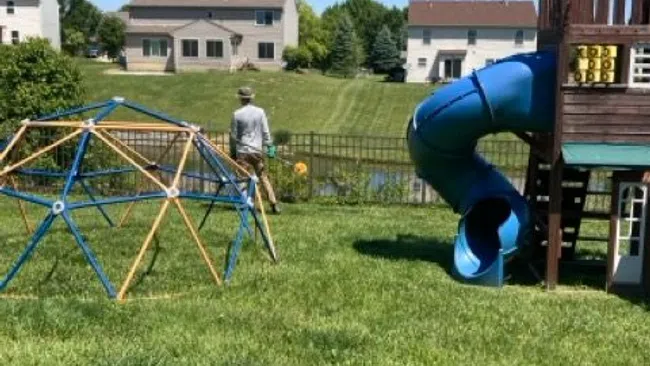 Backyard playground with blue twist slide, climbing dome, and person walking near a pond, suburban houses in background.