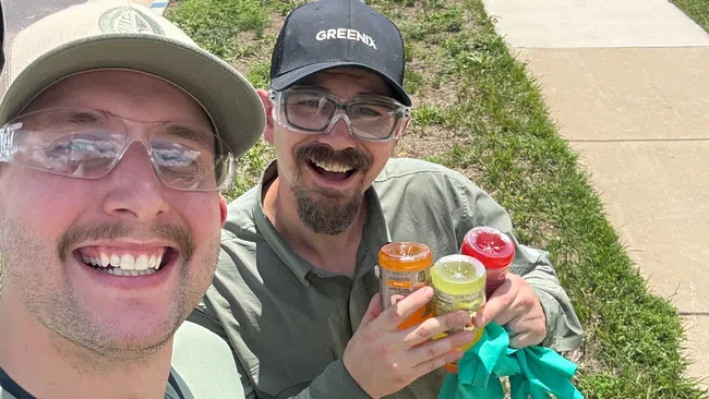 Two men in work uniforms smiling outdoors, one holding containers and gloves, standing on grass by a sidewalk.