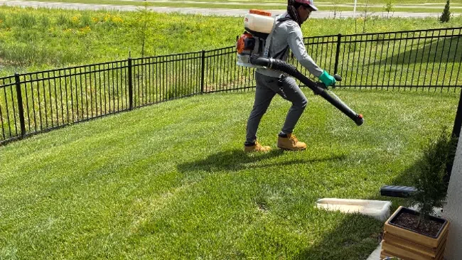 Person wearing gloves and a backpack leaf blower cleaning a green lawn next to a patio with potted plants.