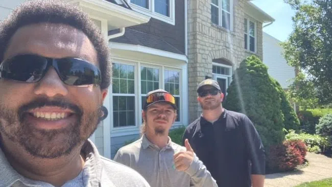 Three men smiling outdoors in front of a suburban house on a sunny day with greenery and clear blue sky.
