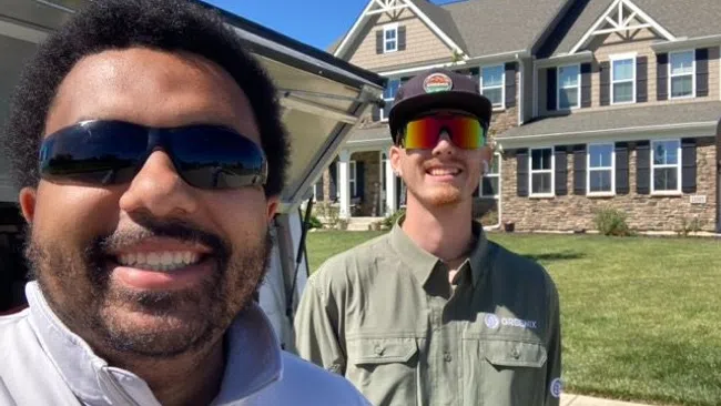 Two men wearing sunglasses smiling outside a large suburban house on a sunny day.
