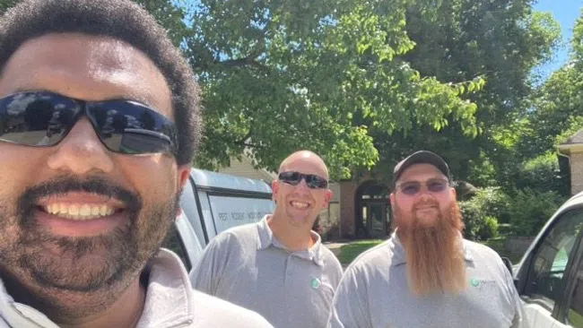Three smiling men wearing Greenix uniforms and sunglasses standing outside near vehicles on a sunny day.