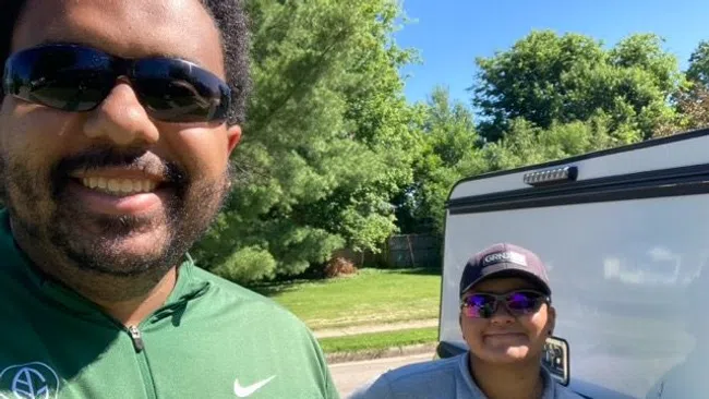 Two smiling people wearing sunglasses outdoors with green trees and a vehicle in the background on a sunny day