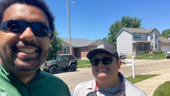 Two smiling people wearing sunglasses and branded shirts outside in a sunny suburban neighborhood.