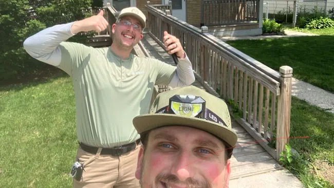 Two smiling men outdoors, one giving thumbs up, standing near a wooden ramp and green lawn in front of a house.