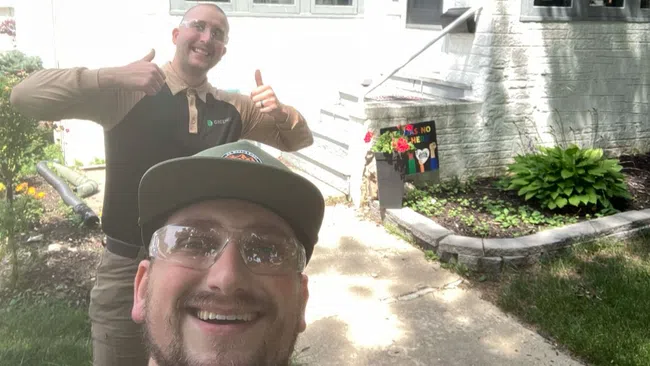 Two men smiling and giving thumbs up outside a white house with garden and steps.