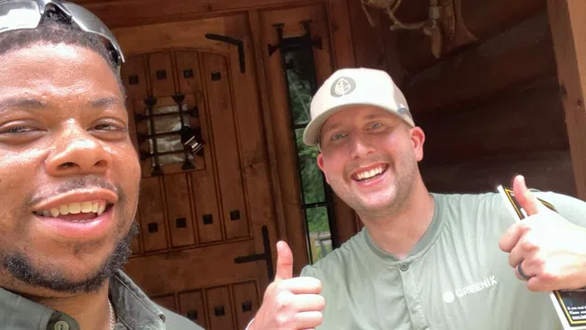 Two men smiling and giving thumbs up outside a wooden cabin entrance with rustic decor.