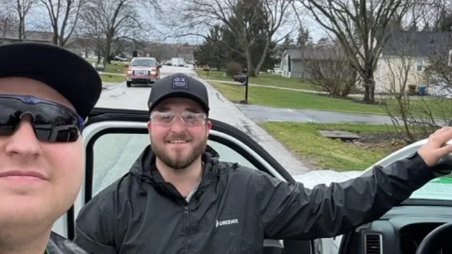 Two men wearing jackets and caps standing by a white vehicle on a cloudy suburban street.