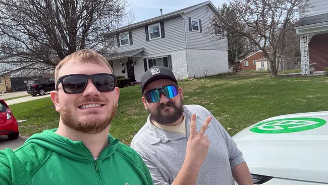 Two men posing outdoors near a white car with green logo in a suburban neighborhood on a cloudy day.