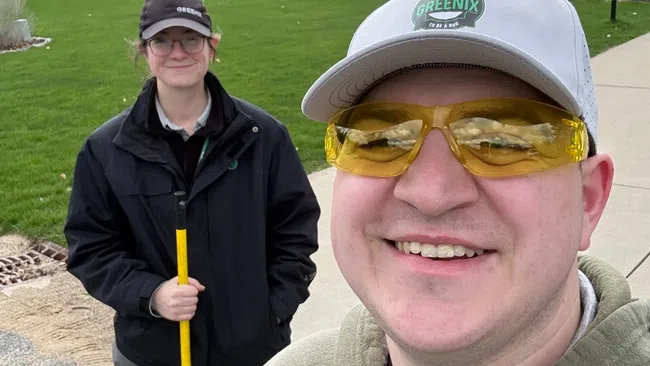 Two smiling landscapers outdoors on a partly cloudy day with green yard and house in the background.