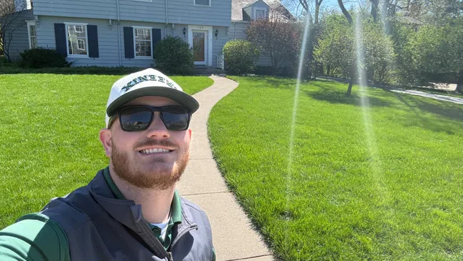 Smiling man in sunglasses and cap taking selfie on sunny day with green lawn and house in background