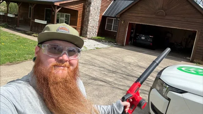 Bearded man wearing safety glasses and cap using a red leaf blower outside a wooden house with a detached garage.