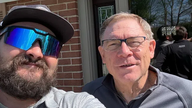 Two men smiling outdoors in front of a brick house with a glass door on a sunny day.