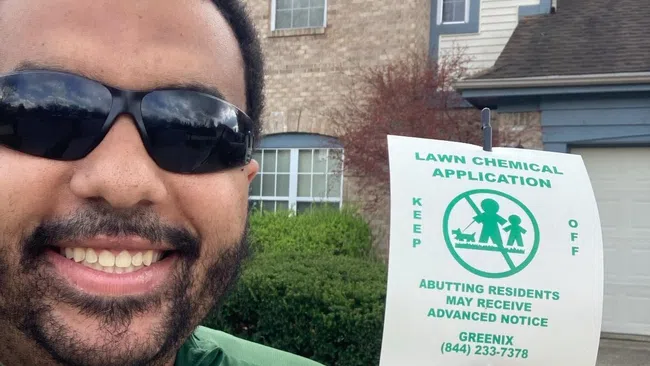 Man in sunglasses smiling while holding home inspection report outside a house with lawn chemical application sign.