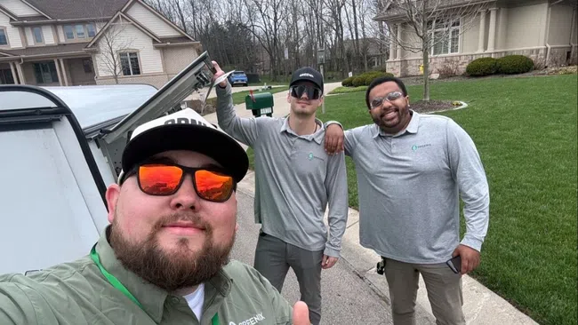 Three men in work uniforms pose outdoors in a suburban neighborhood with thumbs up on a clear day.