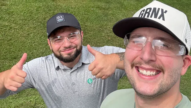 Two smiling men wearing caps and safety glasses give thumbs up on a well-maintained green lawn in front of a house.