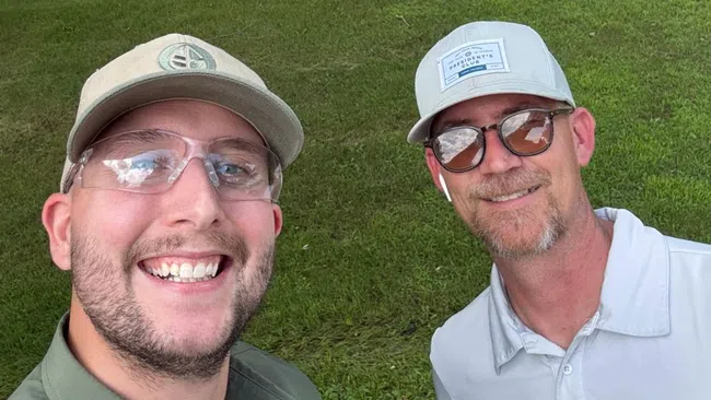 Two men smiling in lawn care uniforms standing on a green grassy yard with trees and houses in the background.