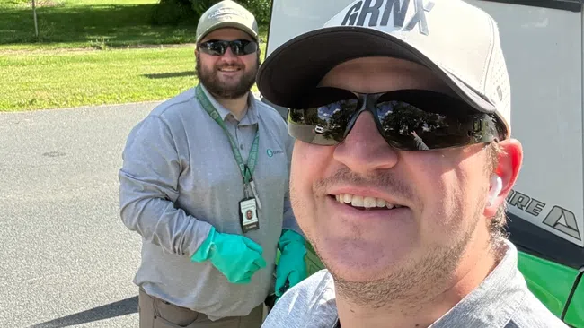 Two men in gray uniforms and sunglasses smiling outdoors near a vehicle and greenery on a sunny day