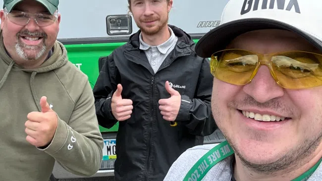 Three smiling men posing outdoors with thumbs up in front of a vehicle on a cloudy day.