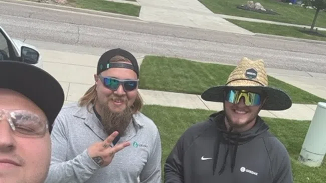 Three men smiling outdoors on a suburban sidewalk, two wearing sunglasses and work attire, one giving thumbs up.
