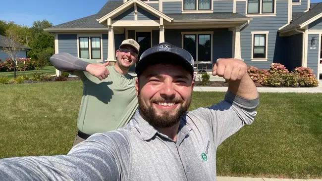 Two men smiling and flexing muscles in front of a large blue house on a sunny day
