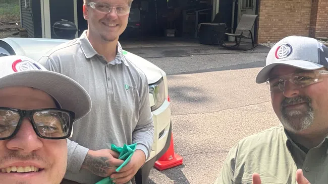Three smiling men wearing work uniforms and safety glasses stand outside a house driveway on a sunny day.