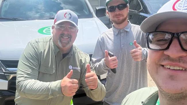 Three men in Green Lawn Service uniforms smiling and giving thumbs up in front of company vehicles in a driveway.