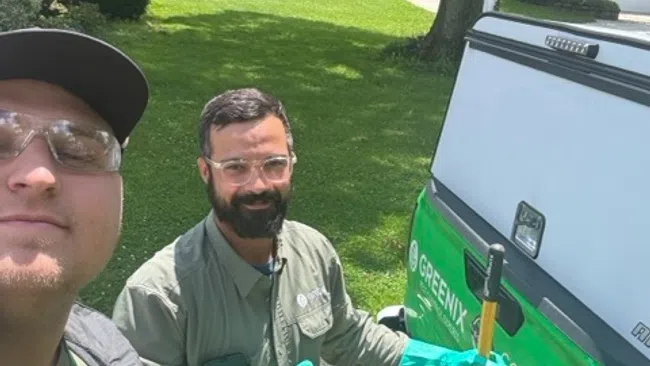 Two pest control workers with gloves and uniforms standing near a Greenix service van outdoors on a sunny day.
