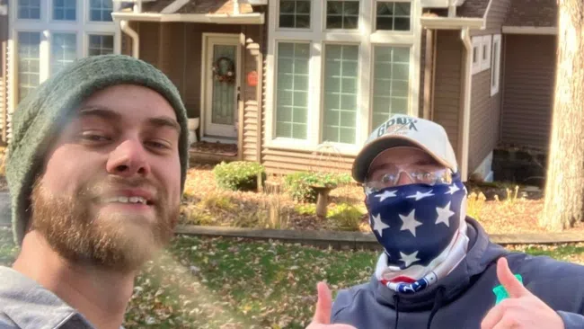 Two men giving thumbs up in front of a brown house with large windows on a sunny autumn day.
