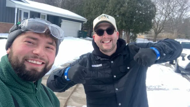 Two men smiling outdoors in winter gear with snow and houses in the background on a cold day.