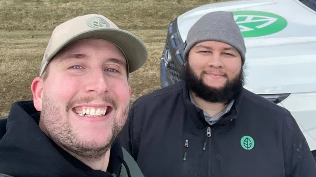 Two smiling men in outdoor jackets and hats stand in front of a large suburban house and a white vehicle with green leaf logo.