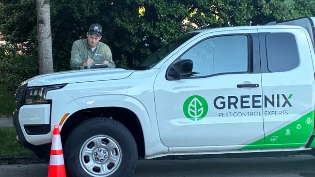 White Greenix pest control truck parked with orange safety cone and technician writing on clipboard outdoors.