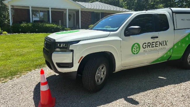 White Greenix pest control truck parked on gravel driveway near house with orange safety cone.